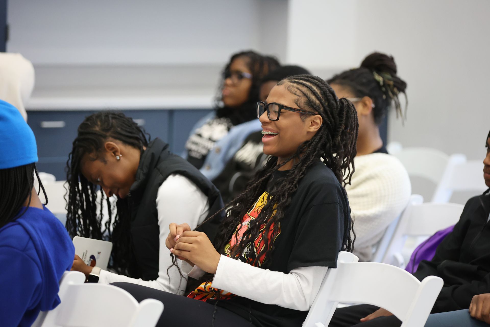 A smiling person in glasses with long braids, sitting in a room with other people.