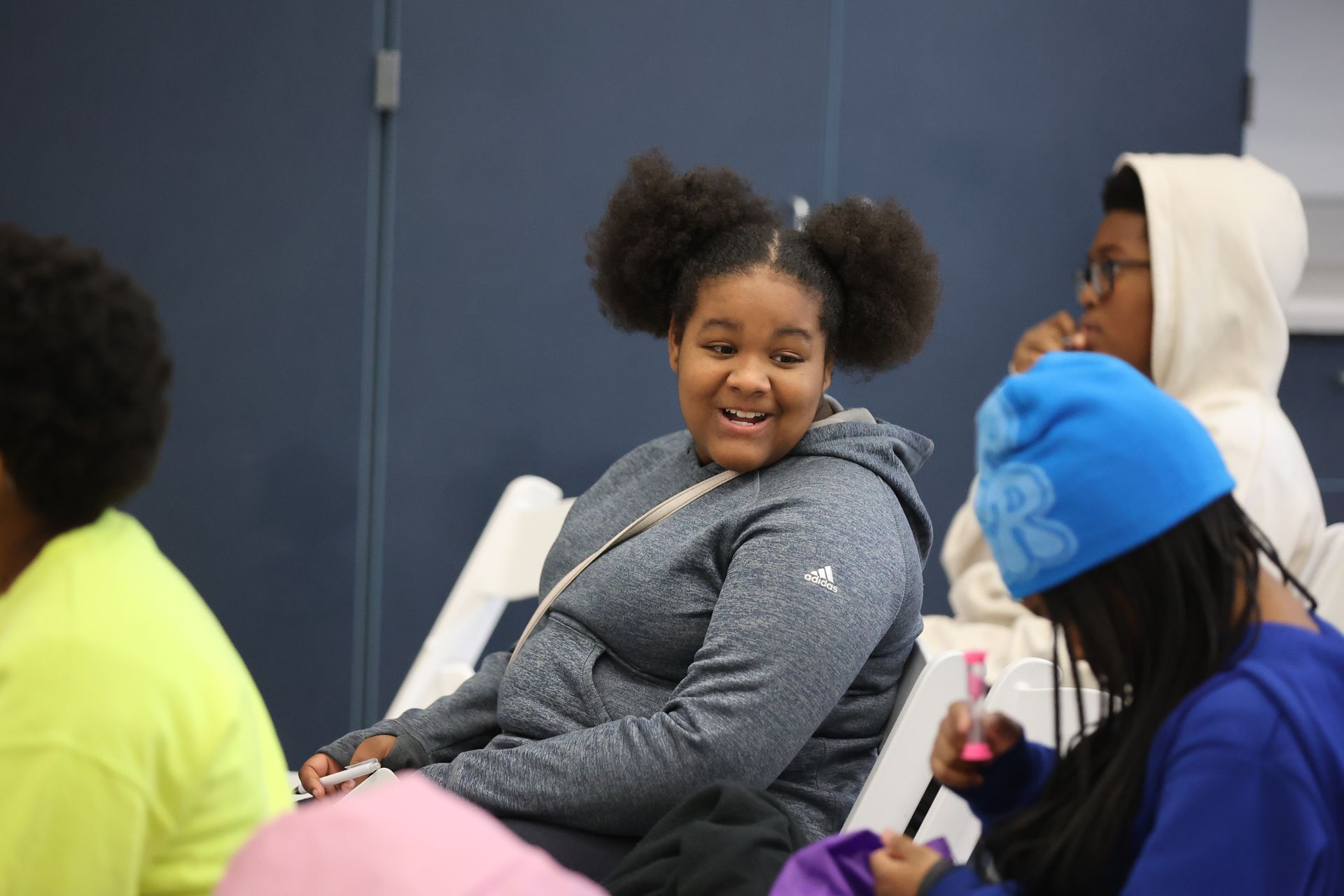 Girl with afro puffs smiles, looking to the side. Others in hoodies, a blue hat in a room.