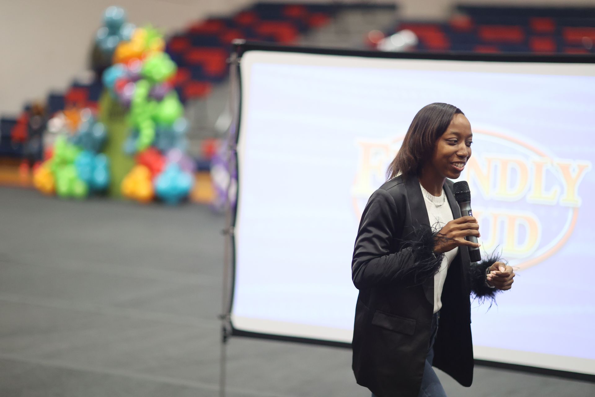 Woman speaking into a microphone in front of a screen with a logo and colorful inflatable toys.