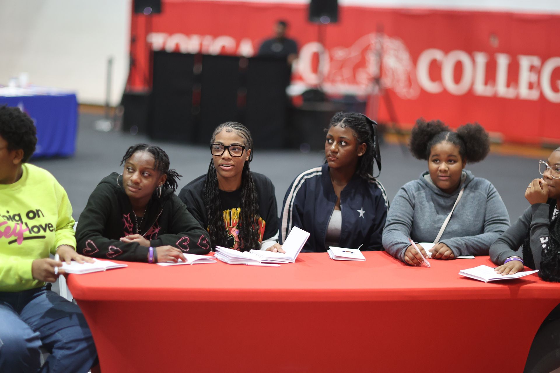 A group of people sit at a red table, some looking forward, in a large space with a