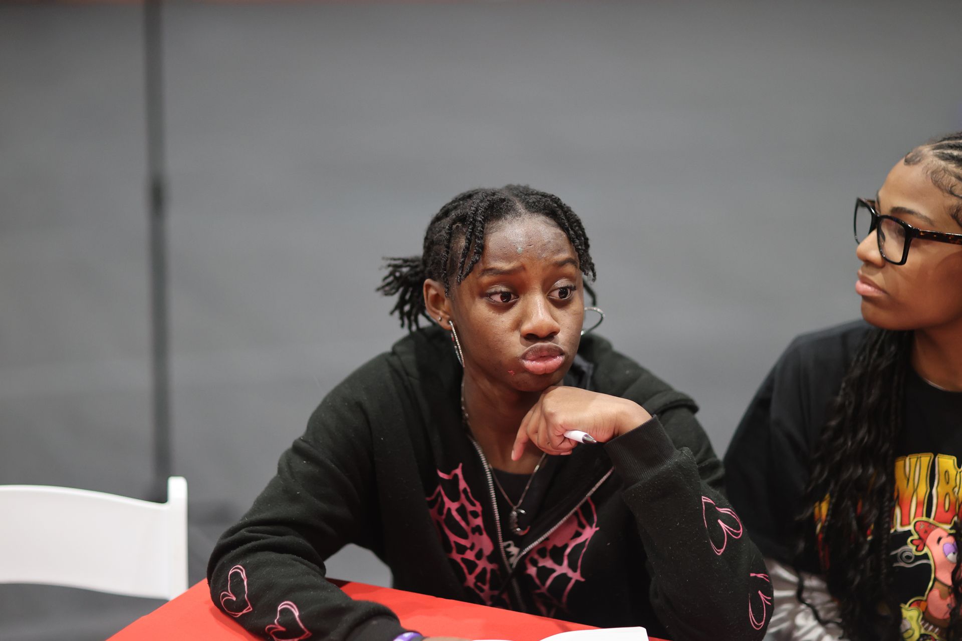 Two people at a table; one looks thoughtful, the other wears glasses and appears focused.
