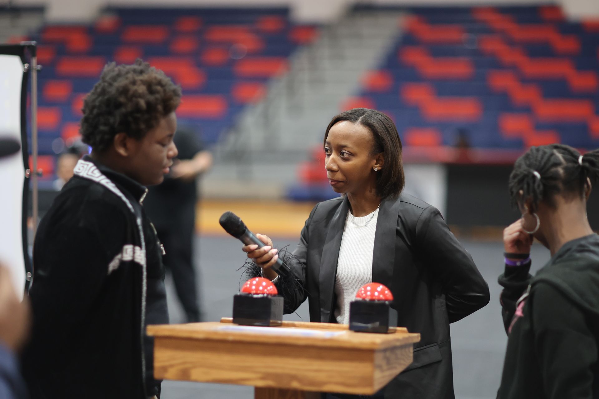 Woman interviewing a person with a microphone near two red buzzers on a podium in an indoor arena.