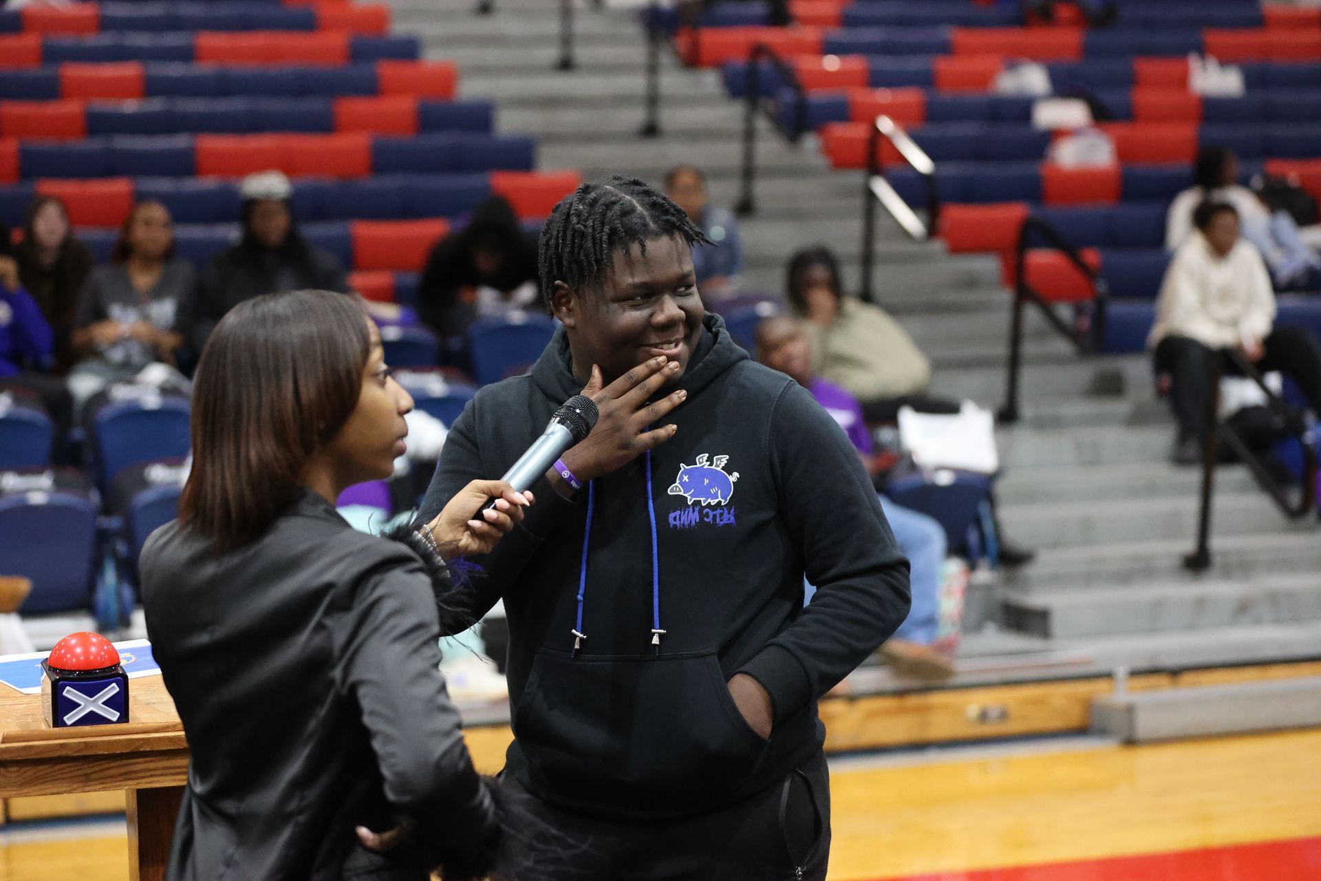 Woman interviewing a person with a microphone in a gymnasium. Audience in bleachers.