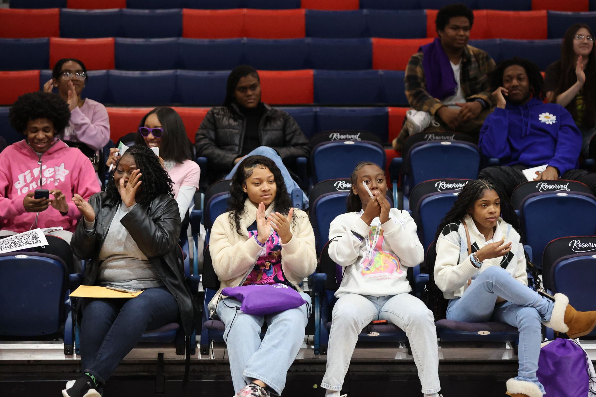 People in bleachers, clapping during an event, some wearing hoodies, jackets, and jeans; blue and red stadium seating.