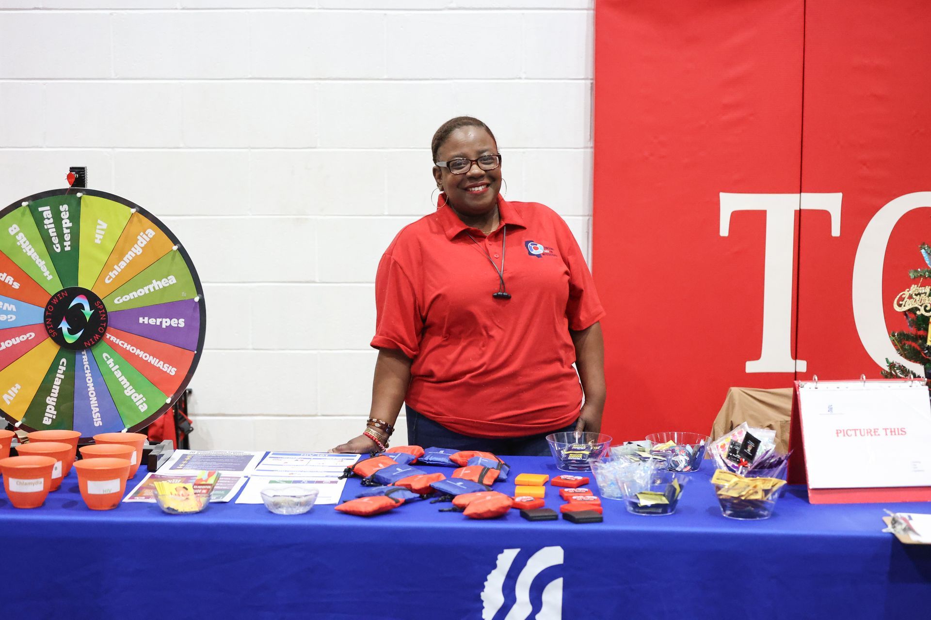 Woman at a table with promotional items, smiling. A prize wheel and red and blue branding are visible.