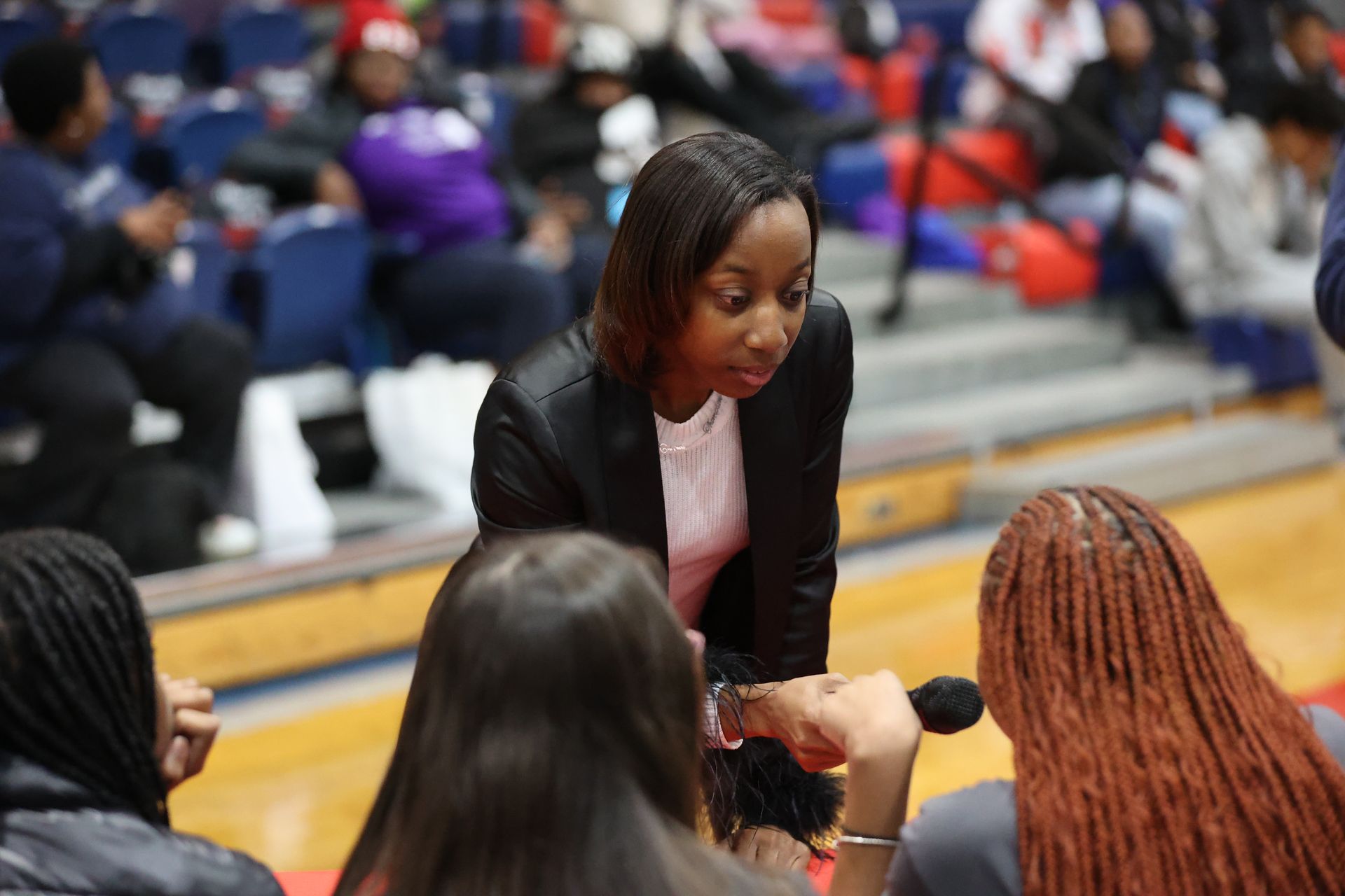 Woman in blazer speaking to a group, holding microphone. Setting appears to be a gymnasium with bleachers.
