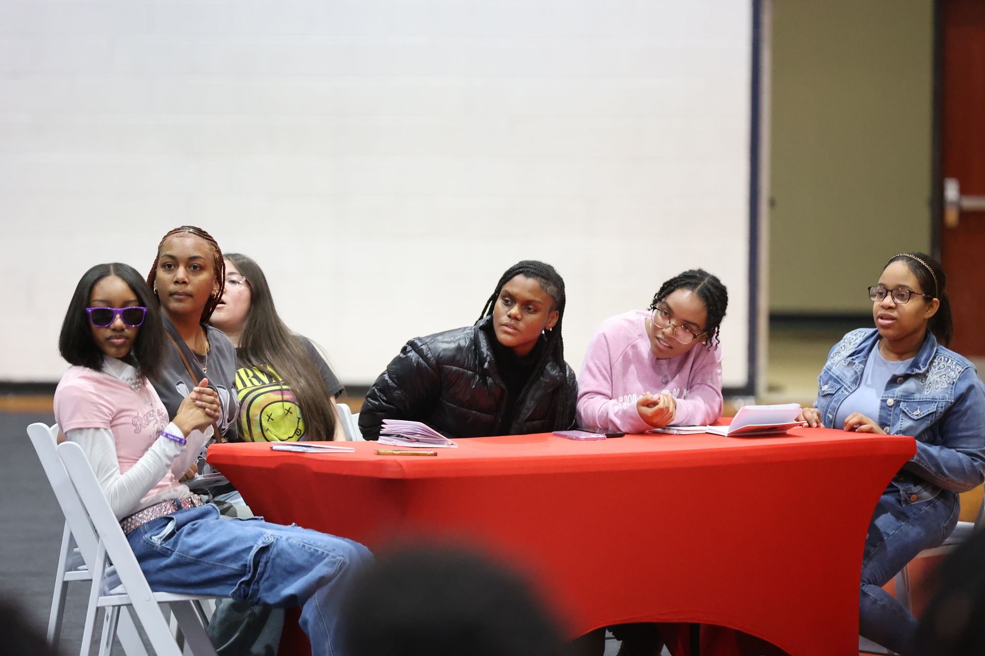 Six people seated at a red table, some looking towards the left, others to the right. A projector screen is in the background.
