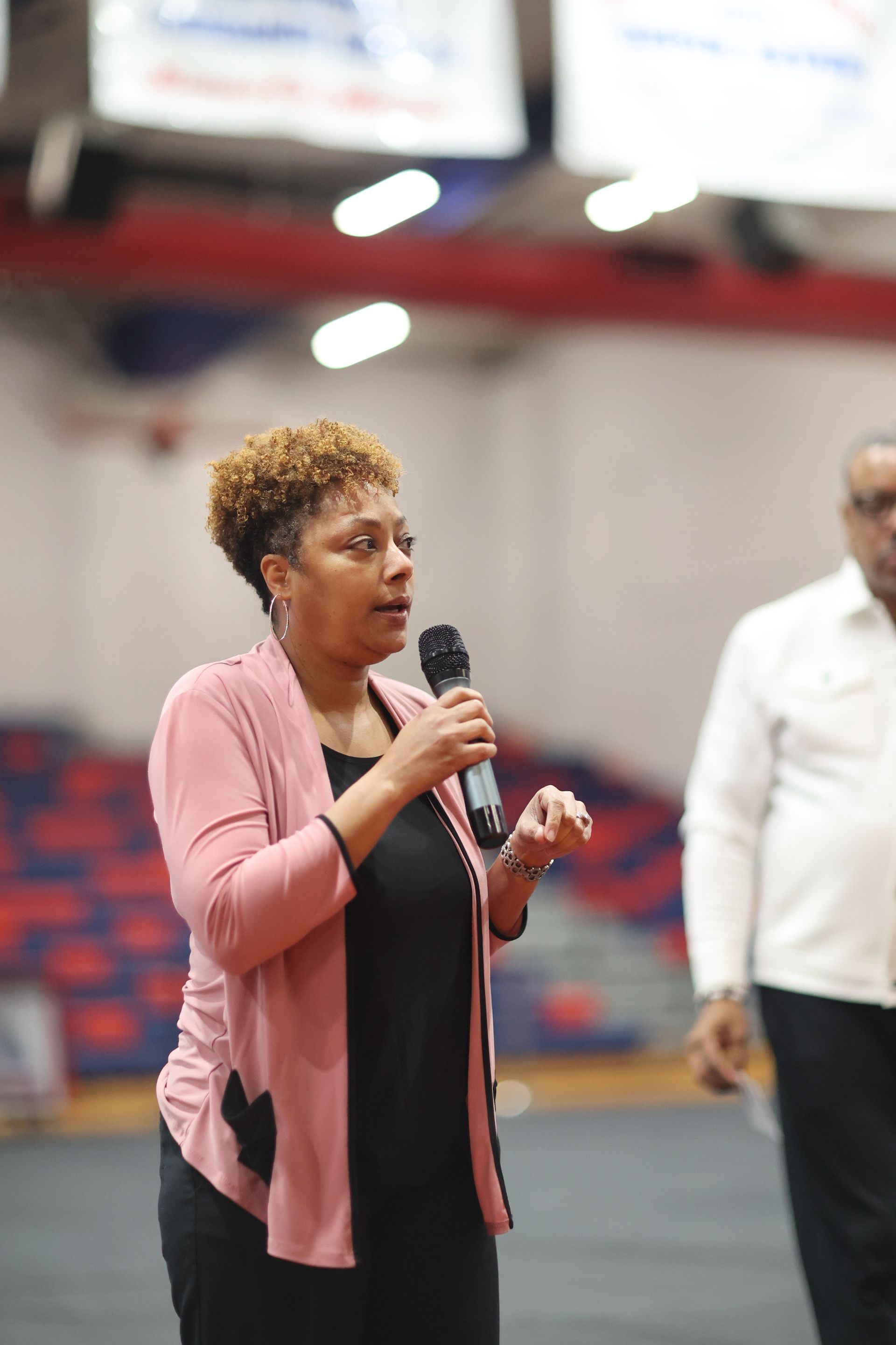 Woman speaking into a microphone in an indoor arena, wearing a pink jacket and black outfit. Another person is in the background. Dr. Melvin