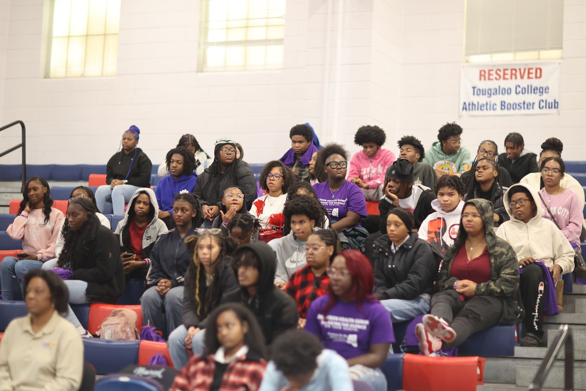 Audience seated in bleachers at an event. Reserved seating sign visible. Blue, white, and red decor.
