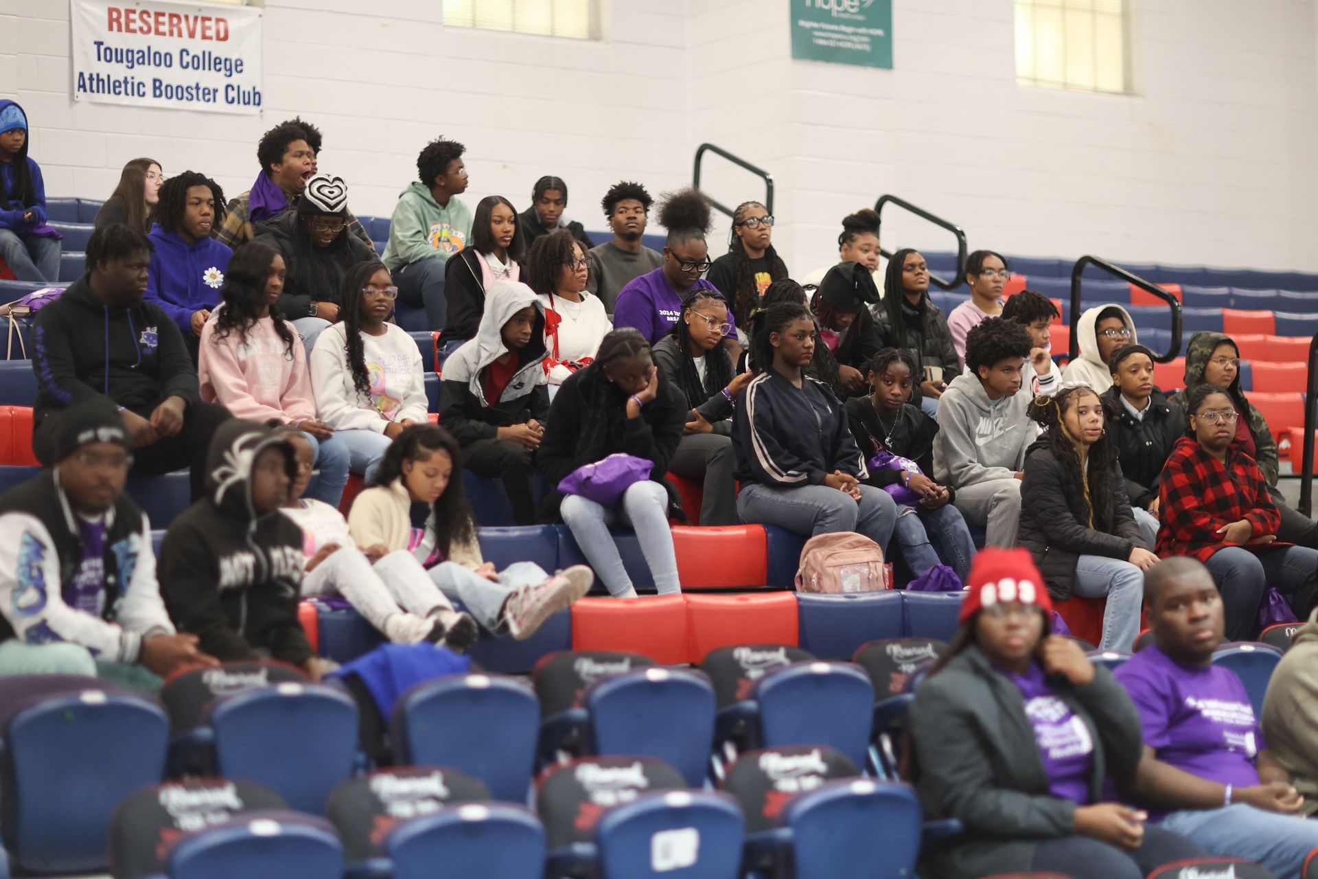 Audience seated in bleachers at an event, many wearing purple. Basketball court and sign visible.