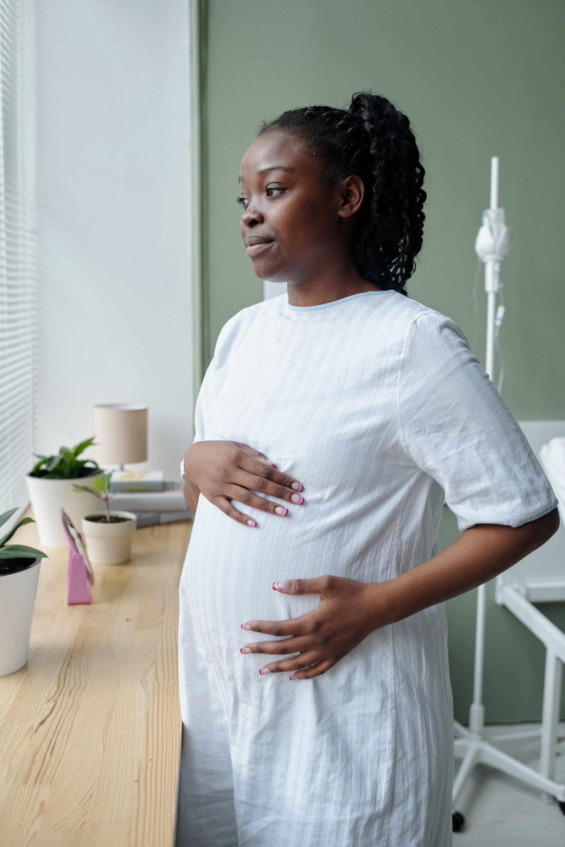 Pregnant person in a white dress, touching her belly, looking out a window.