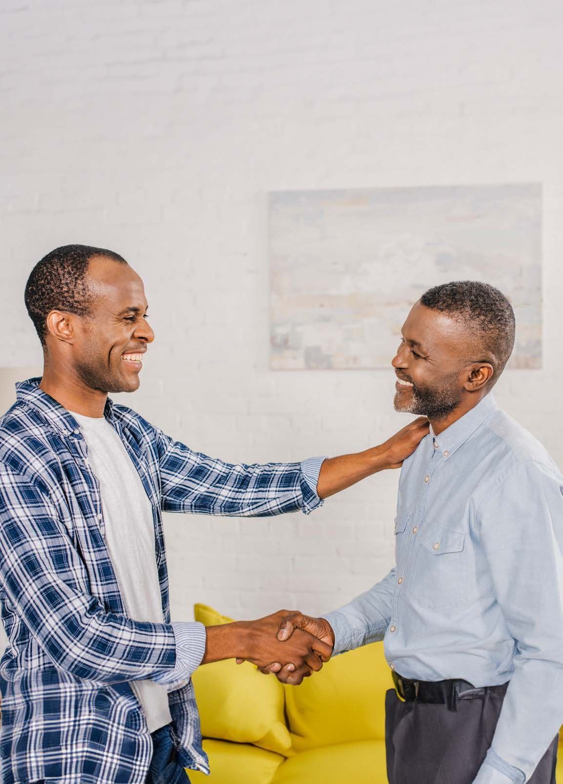 Two people shaking hands and smiling in a home setting. One person has their hand on the other's shoulder.