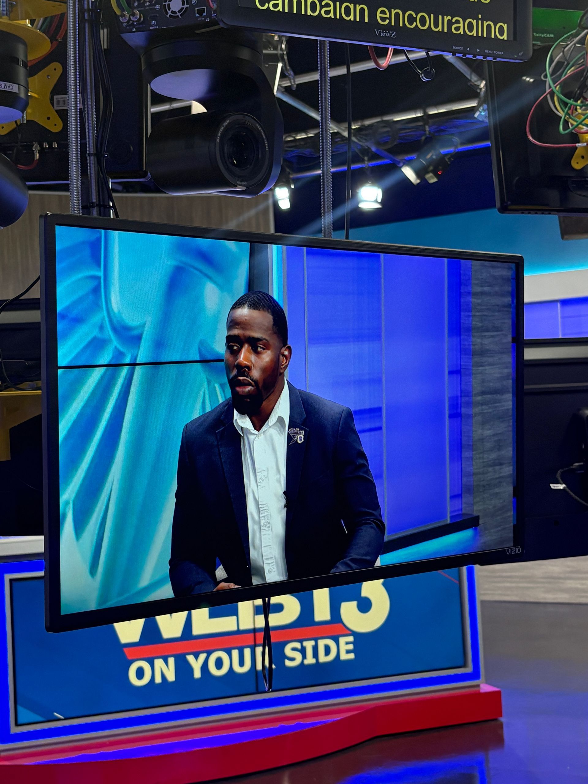 Man in a suit on a news set; on-screen background is blue; news channel graphic is visible.