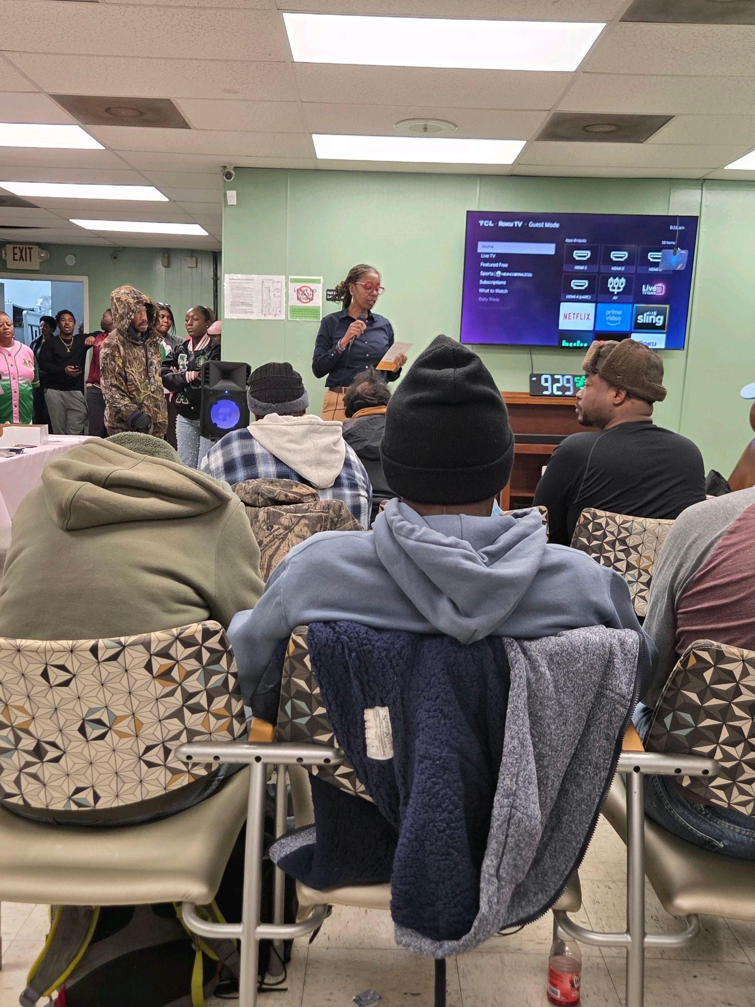People listen to a woman speaking at a meeting, in a room with a TV.
