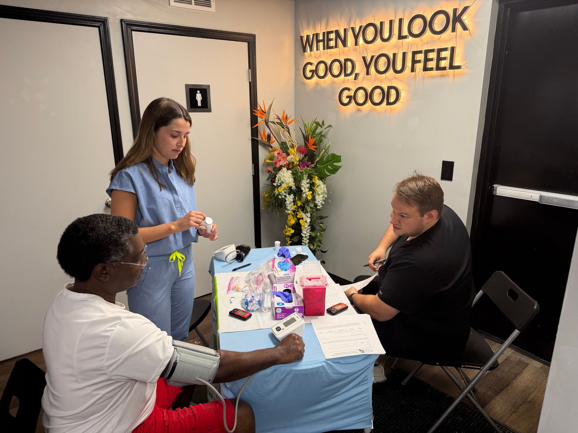 Three people at a table during a health fair, with flowers and a lit sign reading “WHEN YOU LOOK GOOD, YOU FEEL GOOD.”