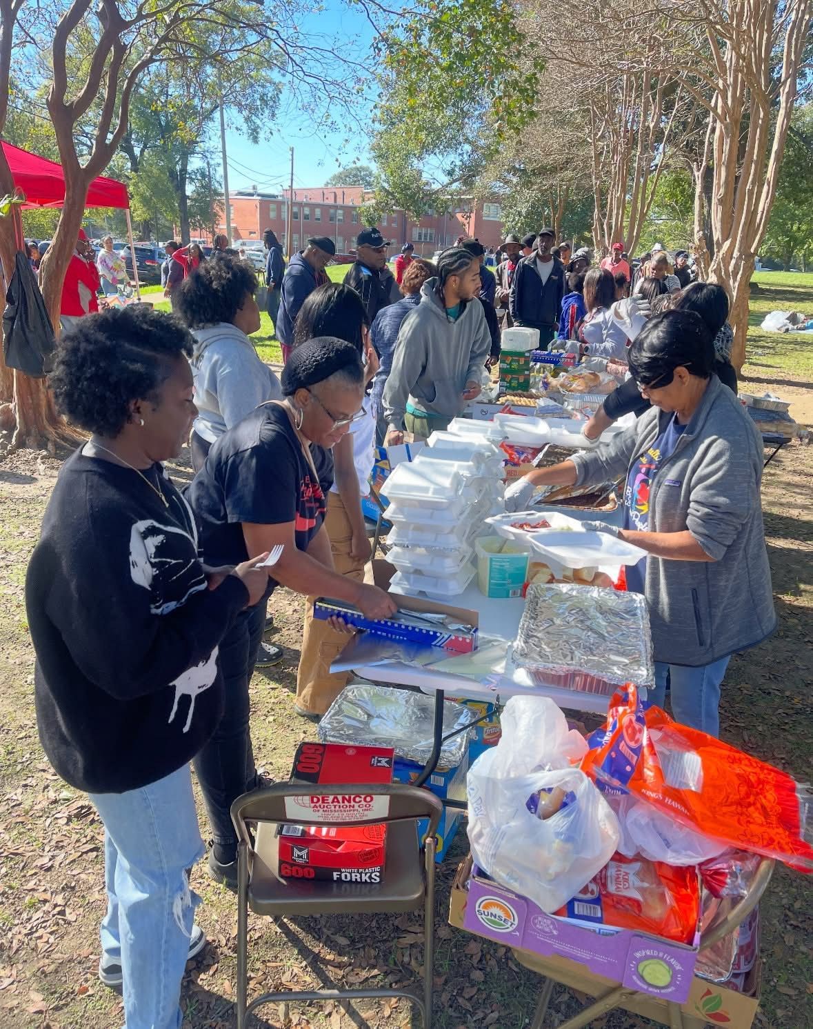 People serving food at an outdoor event on a sunny day. Food on tables, people in line.