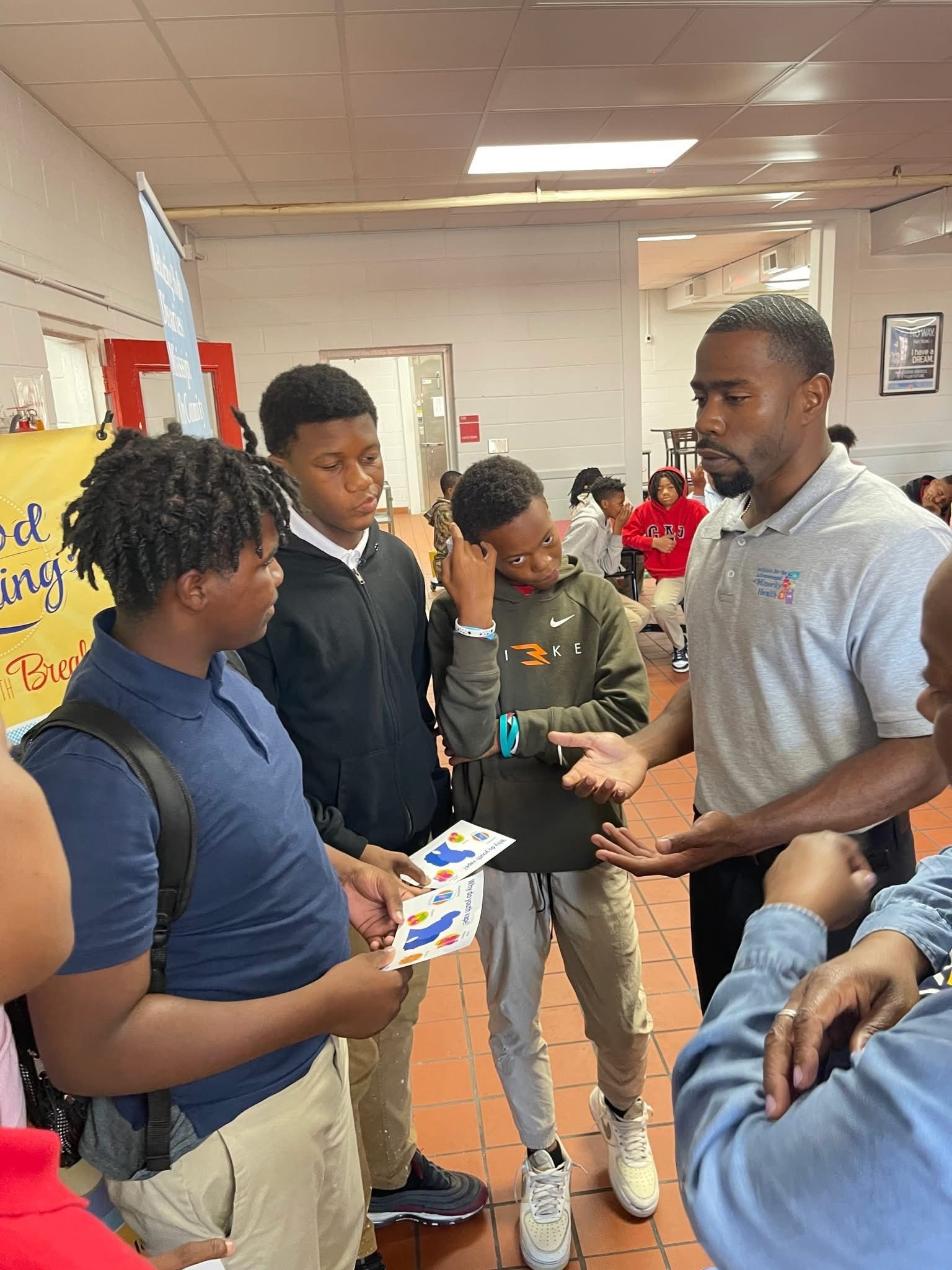 A man talks to three students, holding cards, in a school hallway.