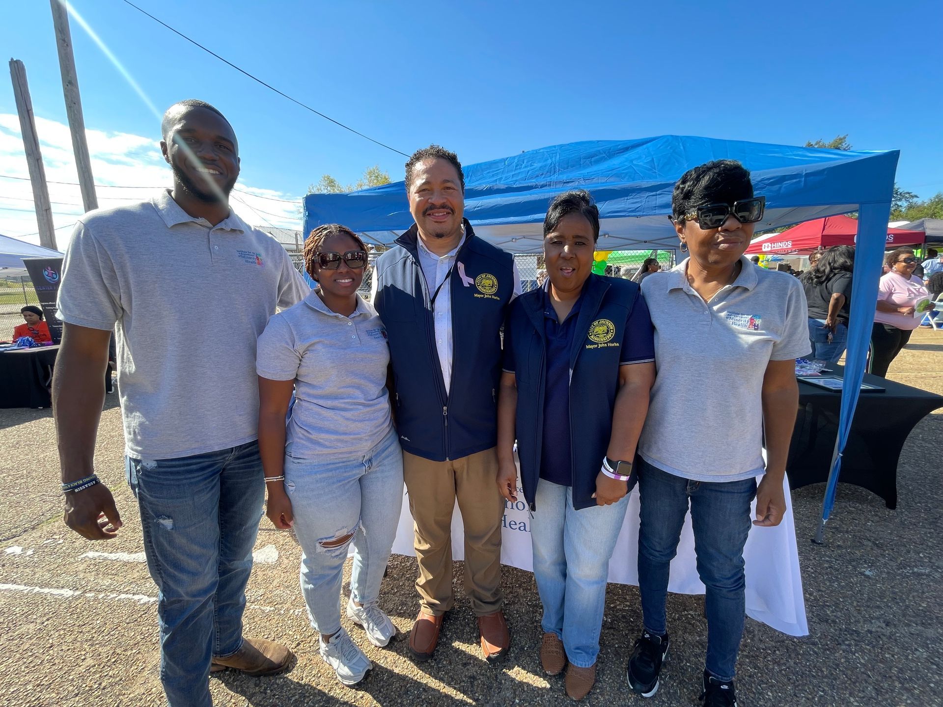 Five people pose outdoors; event tent in background.