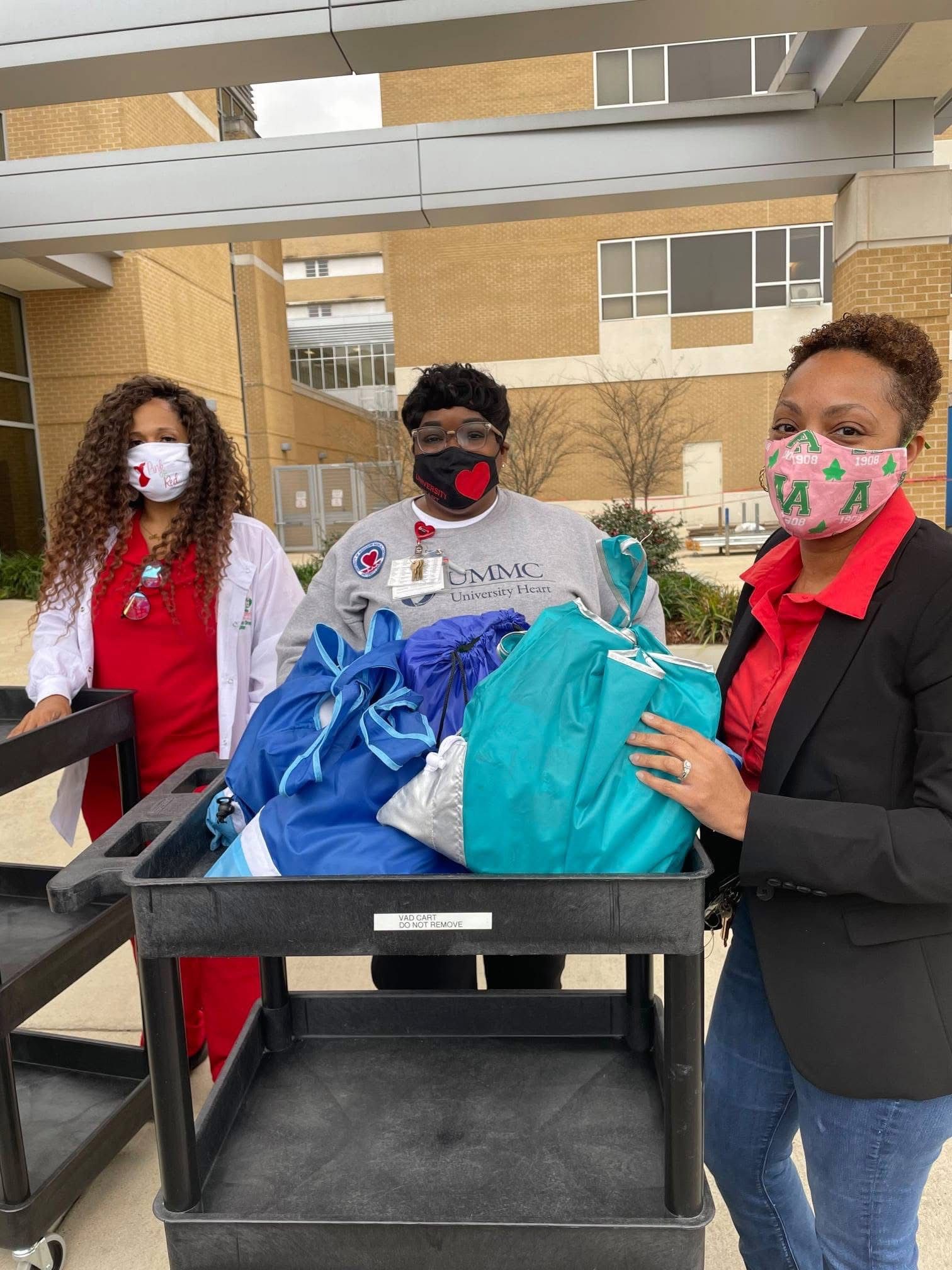 Three people with masks stand by a cart filled with blue bags outdoors.
