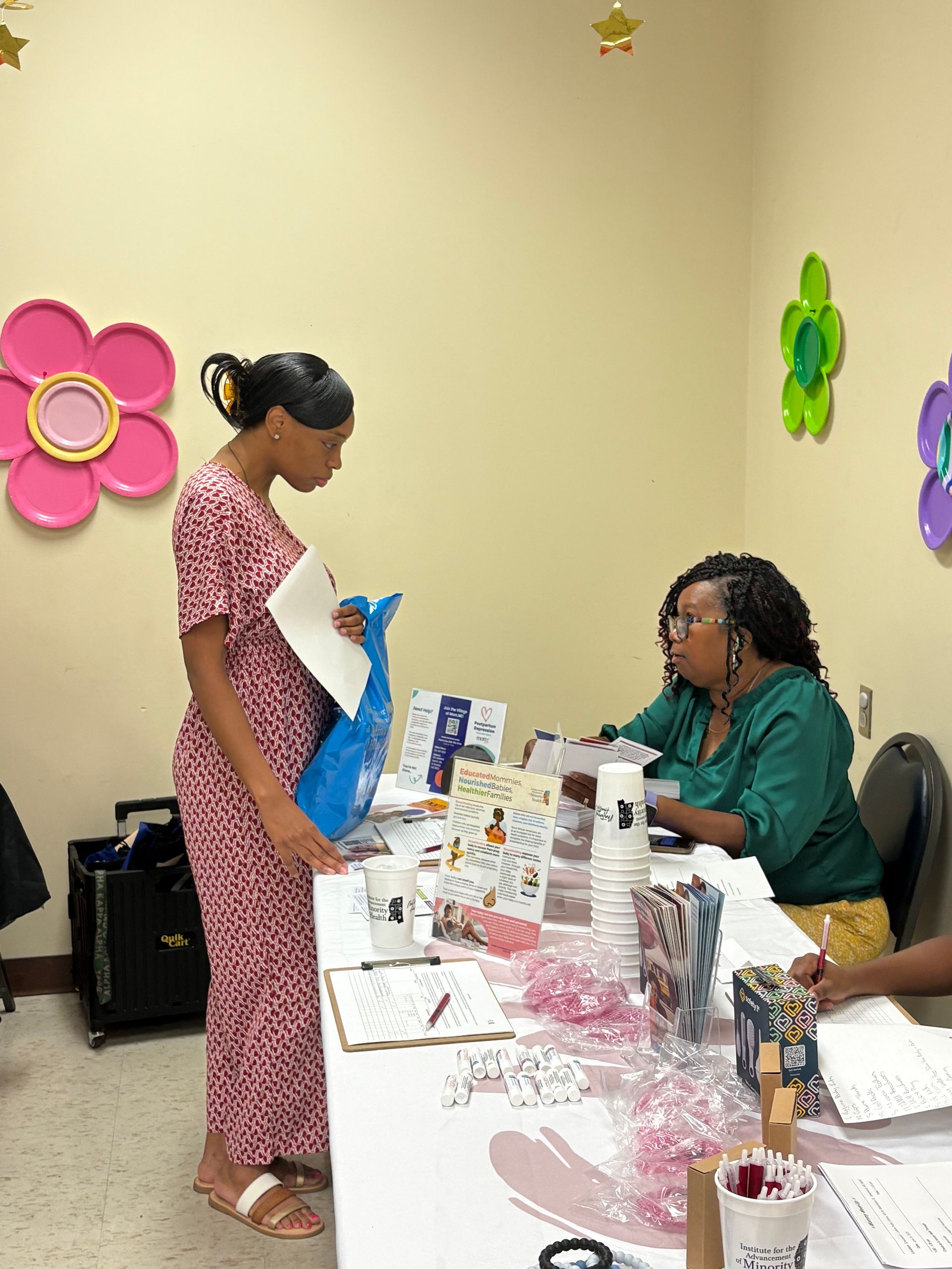 Woman at a desk, speaking to another woman in a red print dress, holding a blue gift bag.