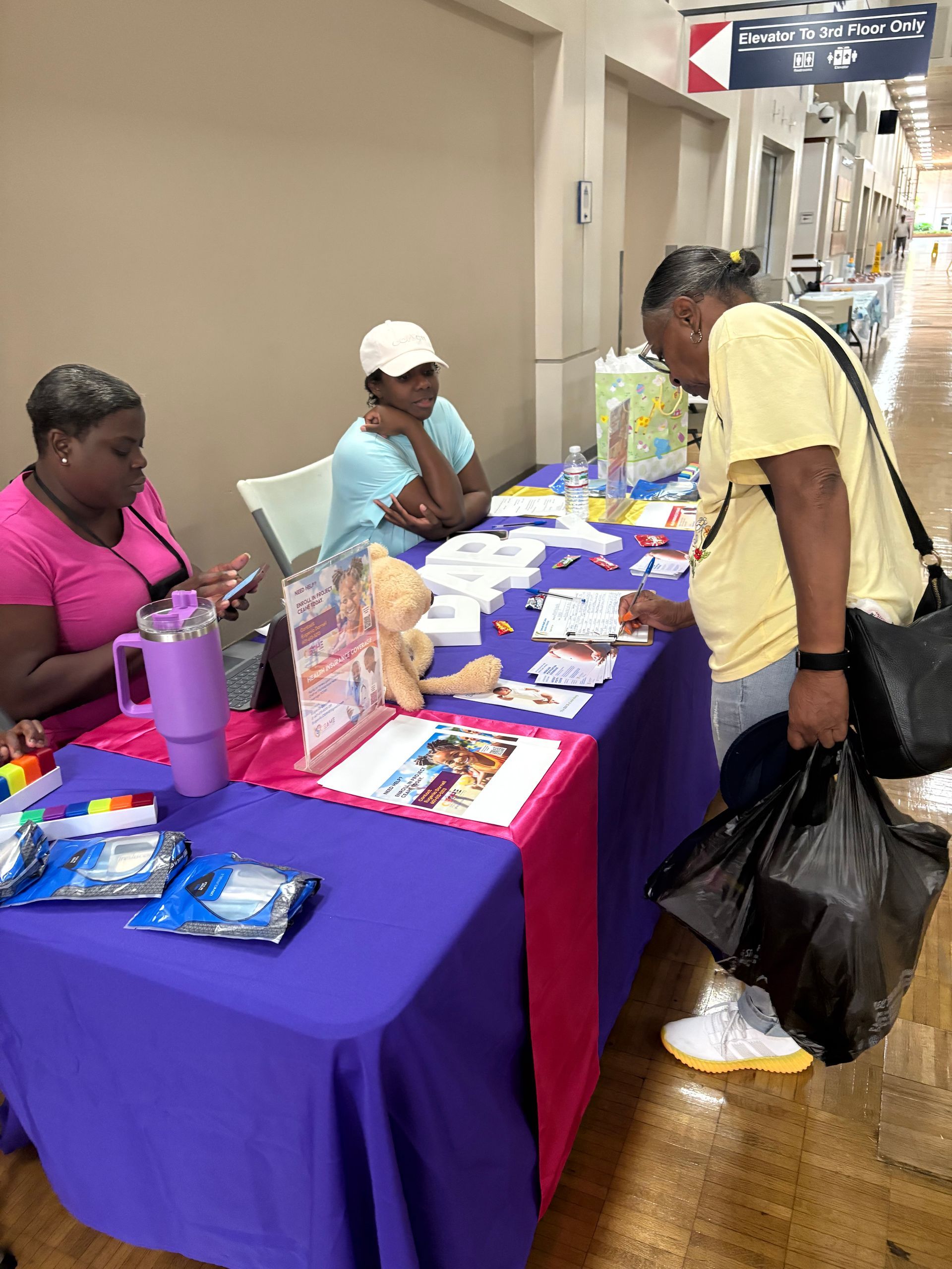 People at a table with brochures and items. One woman leans over table, two women sit behind it. The table has a purple and pink tablecloth.