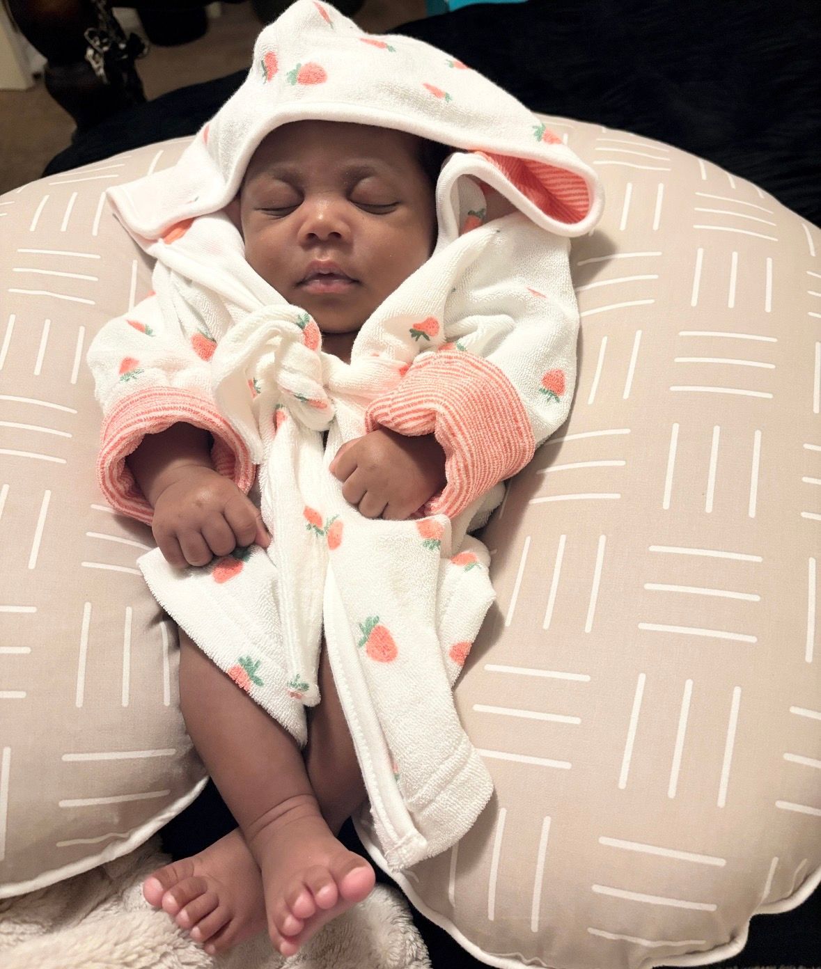Sleeping baby in a white hooded robe, resting on a beige nursing pillow with pink accents. IAMH Maternal and Child Health.