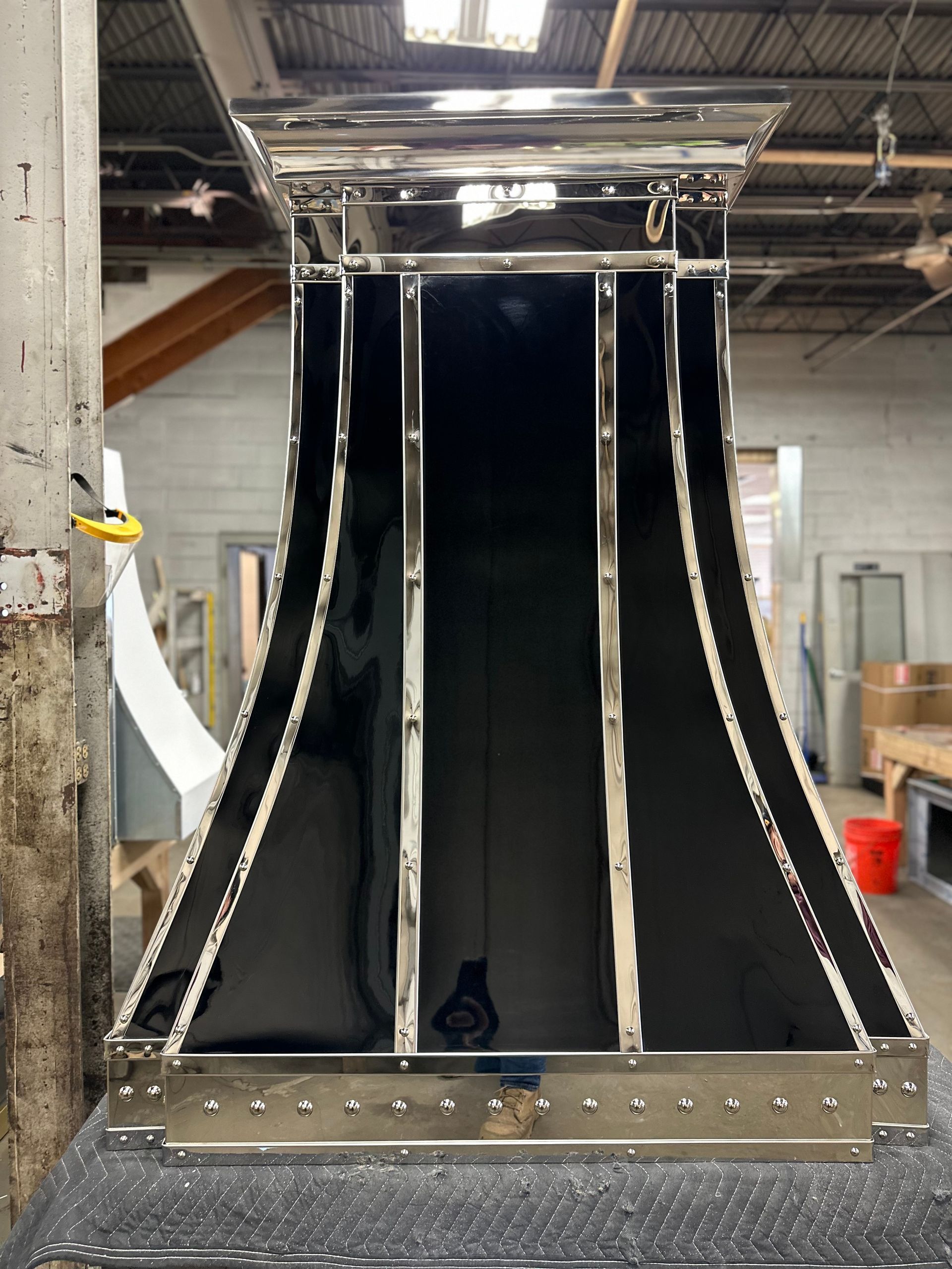Black glossy industrial chute or funnel in a workshop, framed by metal rails and overhead machinery.