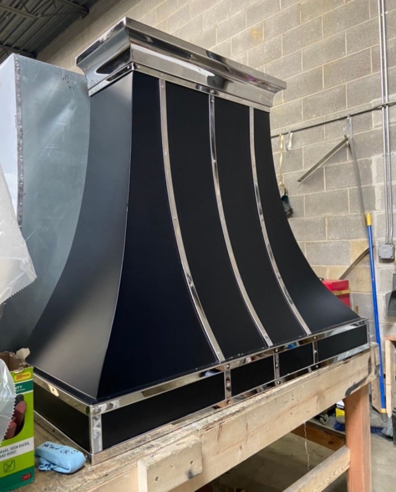 Large black metal duct or housing on a workbench in a workshop, with cinder block walls behind it.