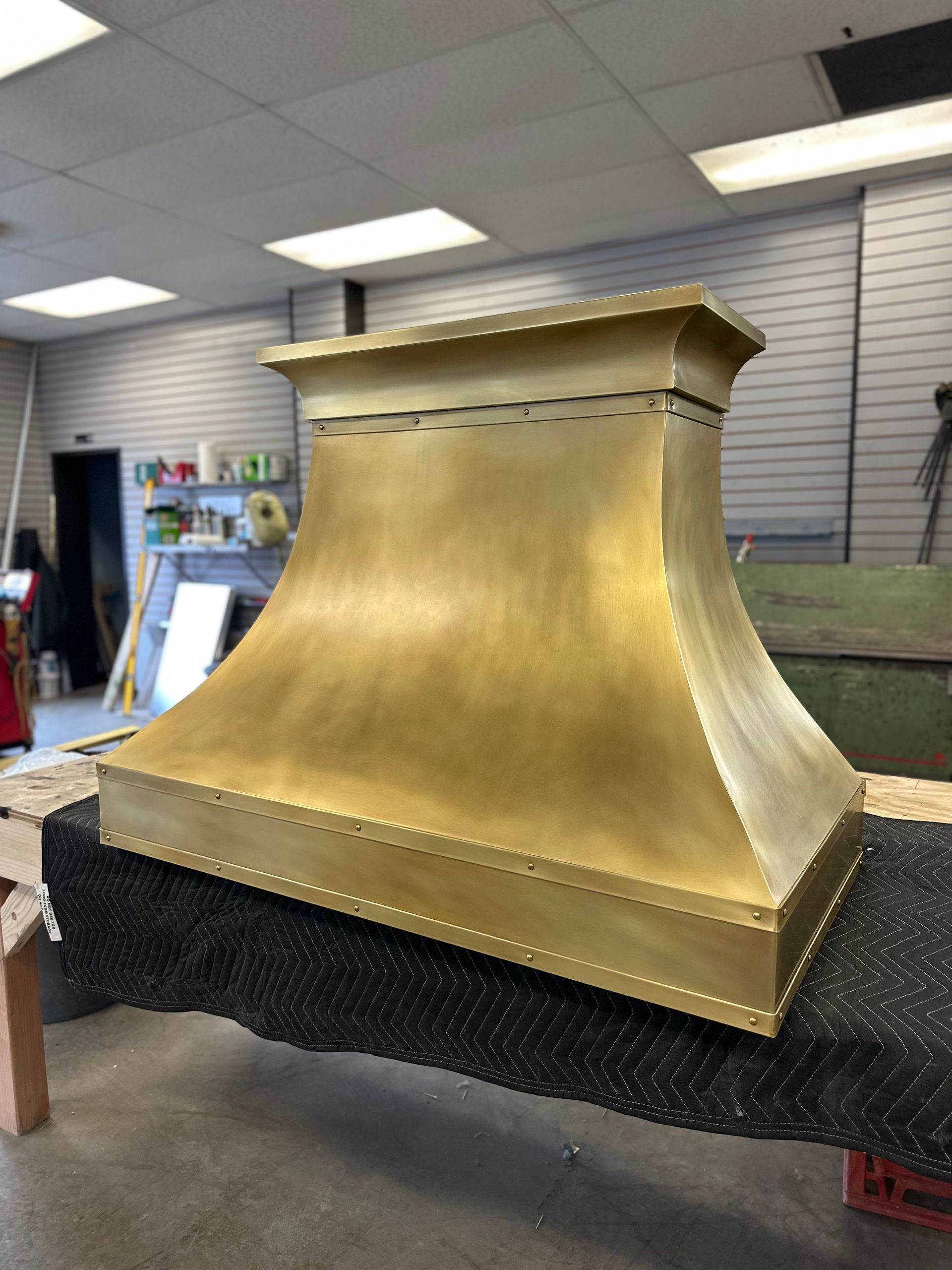 Gold metal hood or chimney cap on a workbench in a workshop, with black foam beneath it.