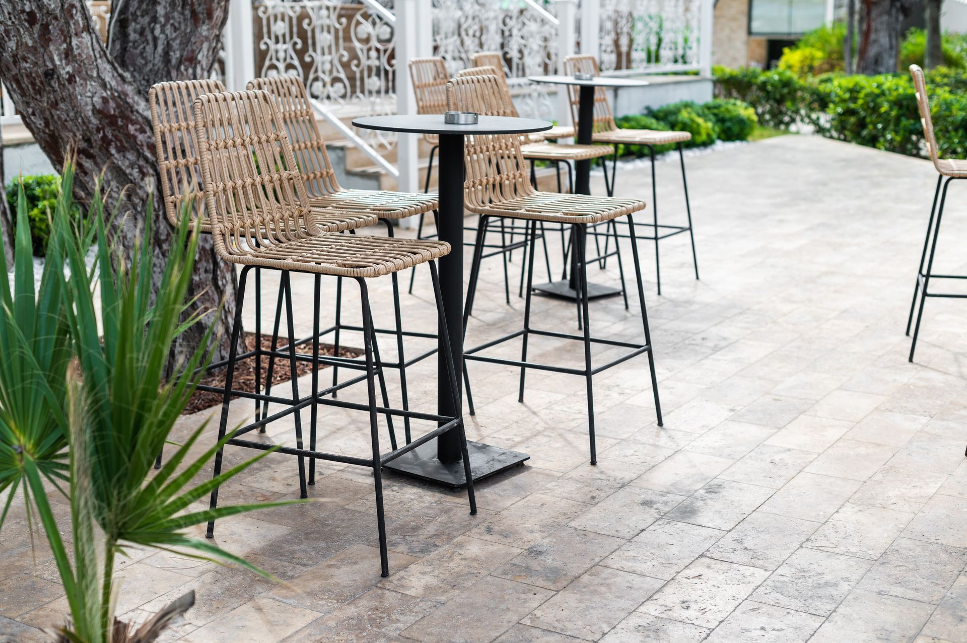 Outdoor café seating with wicker bar stools and round tables on a stone patio near greenery