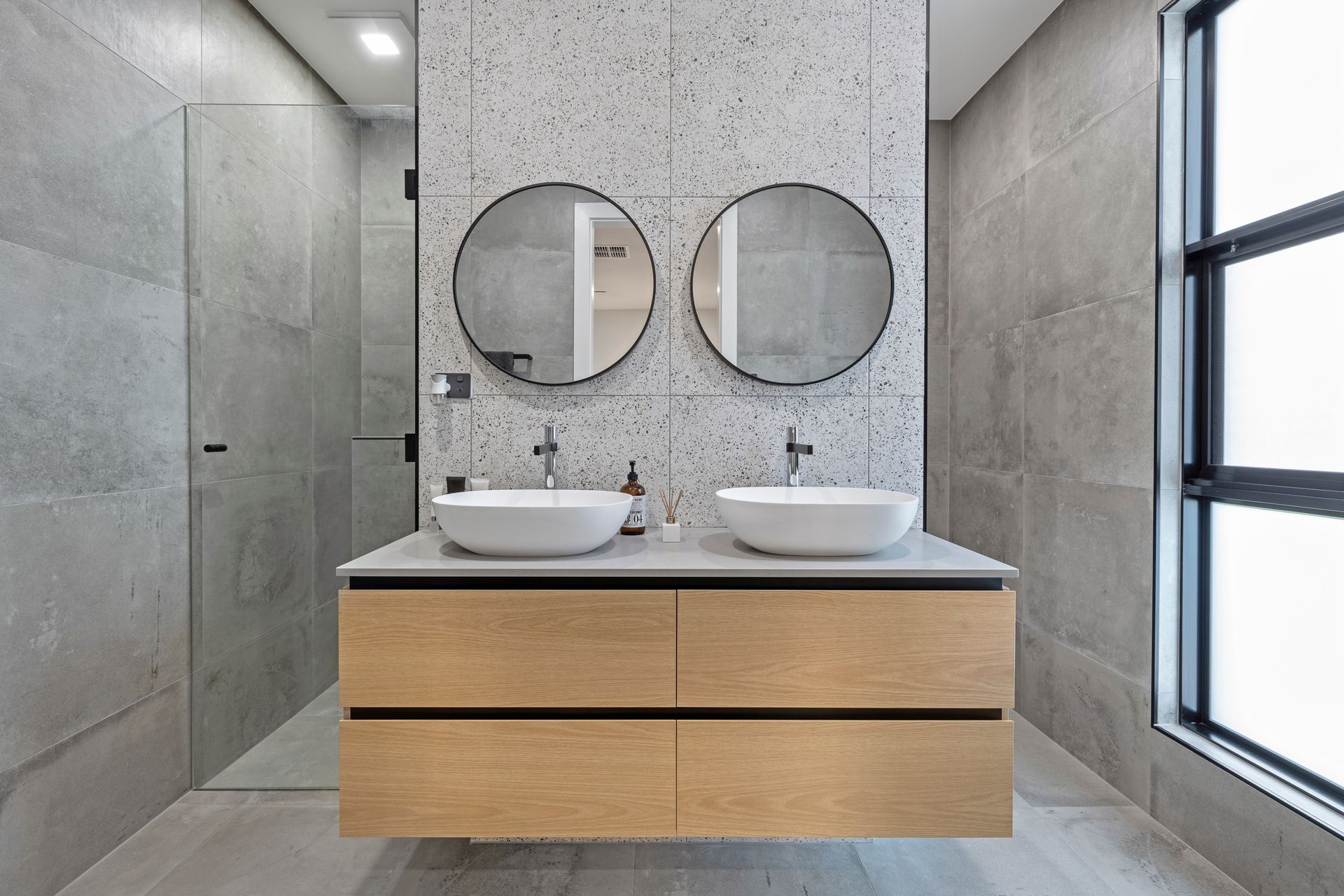 Modern bathroom with double sinks, round mirrors, gray tile, and a large window