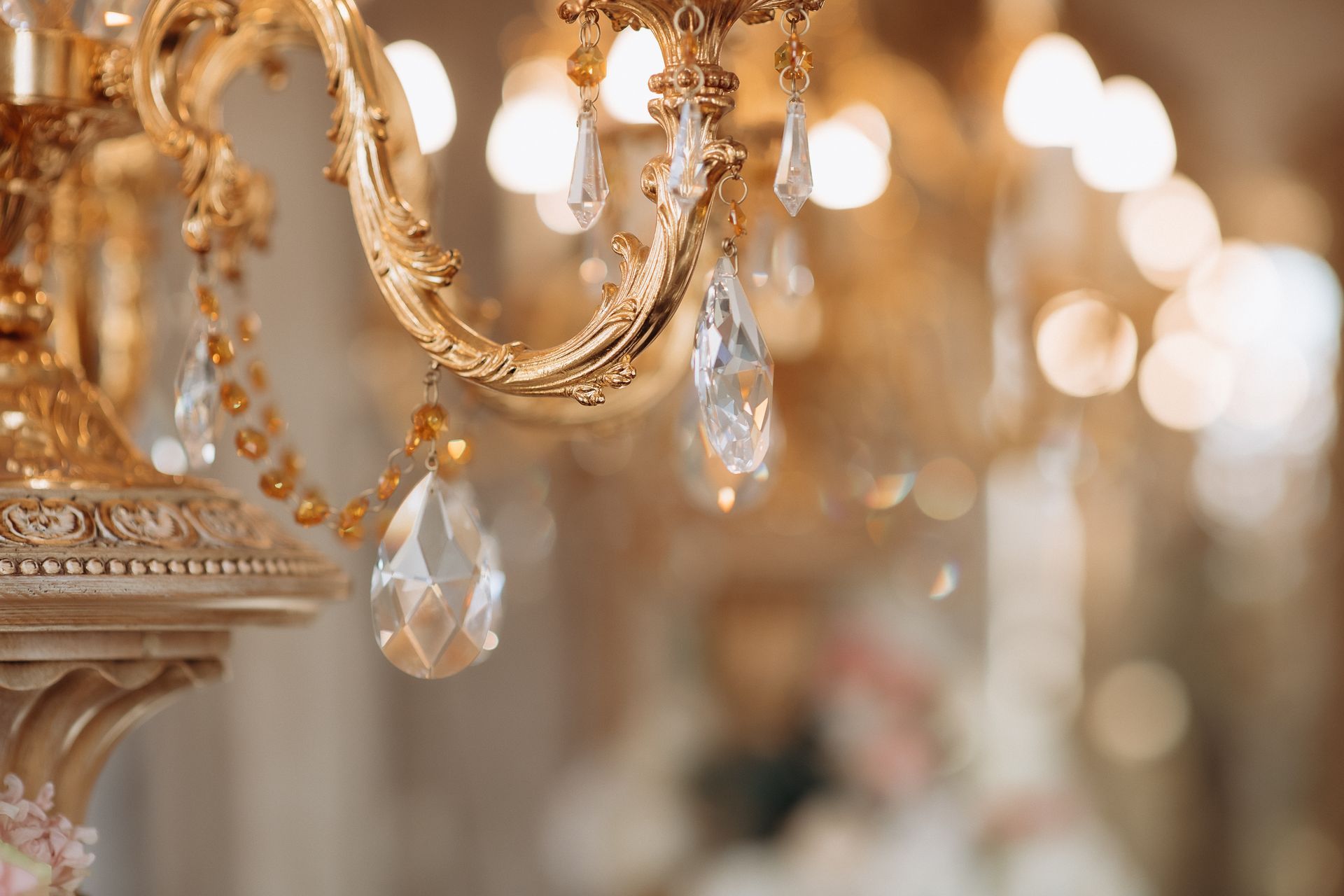 Ornate gold chandelier with hanging crystal drops and warm blurred lights in the background