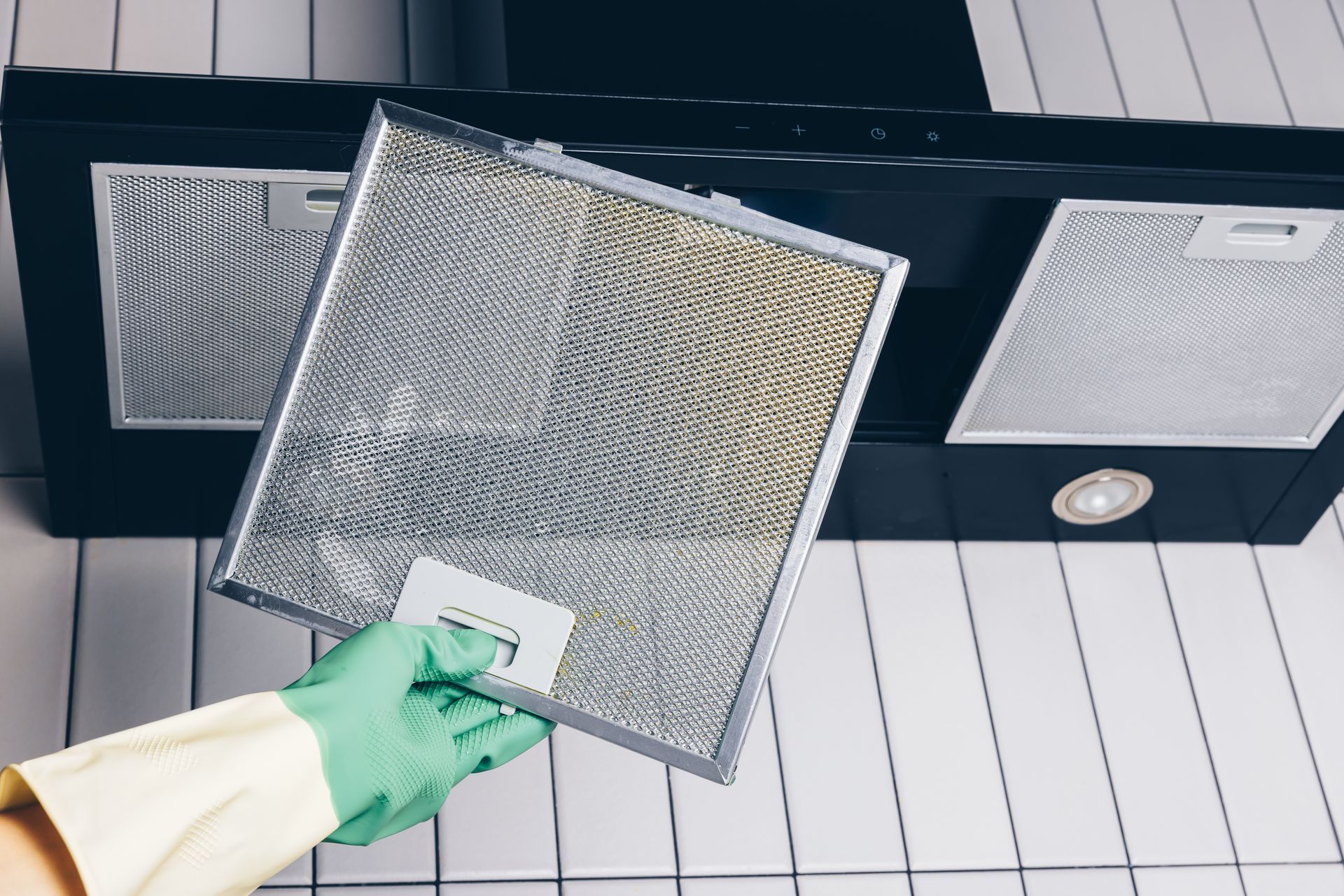 Hand wearing green glove removing a metal kitchen range hood filter underneath the vent.