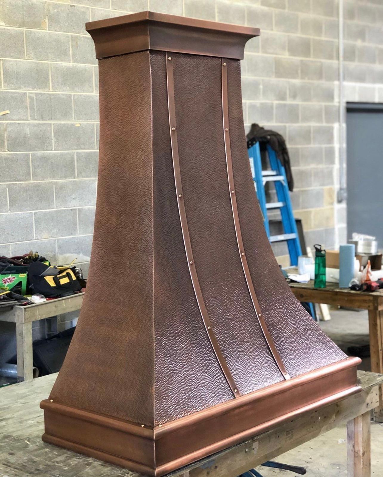 Copper range hood in a workshop, with a textured curved body and wood trim on a bench