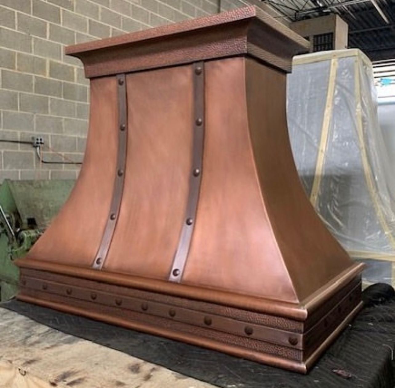 Large copper range hood with dark metal accents in a workshop or storage area