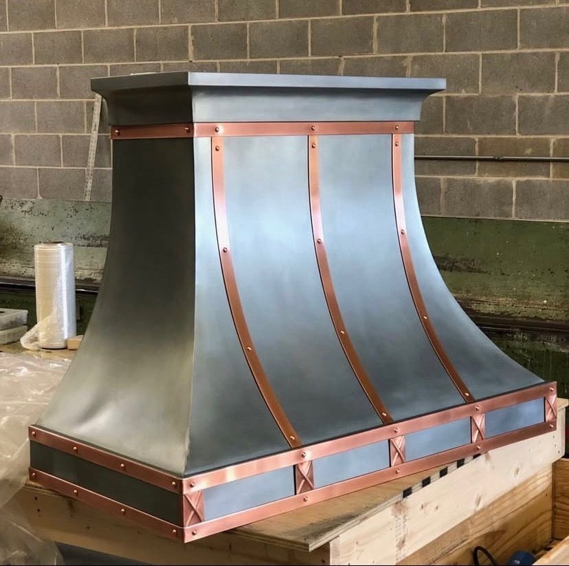 Copper and black metal range hood in a workshop, with curved panels and a wooden base.