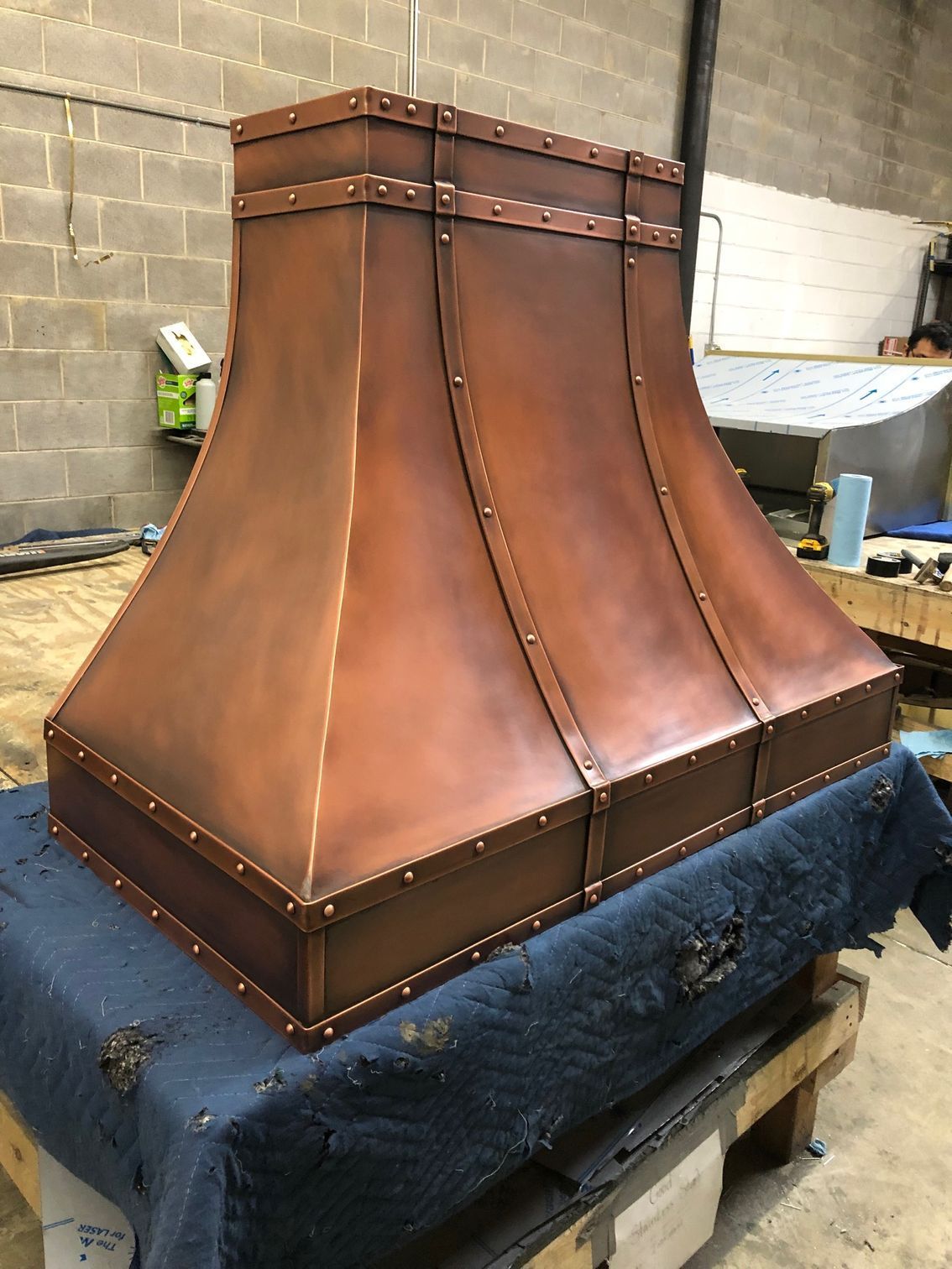 Copper range hood on a workbench in a workshop, with riveted panels and a square top.