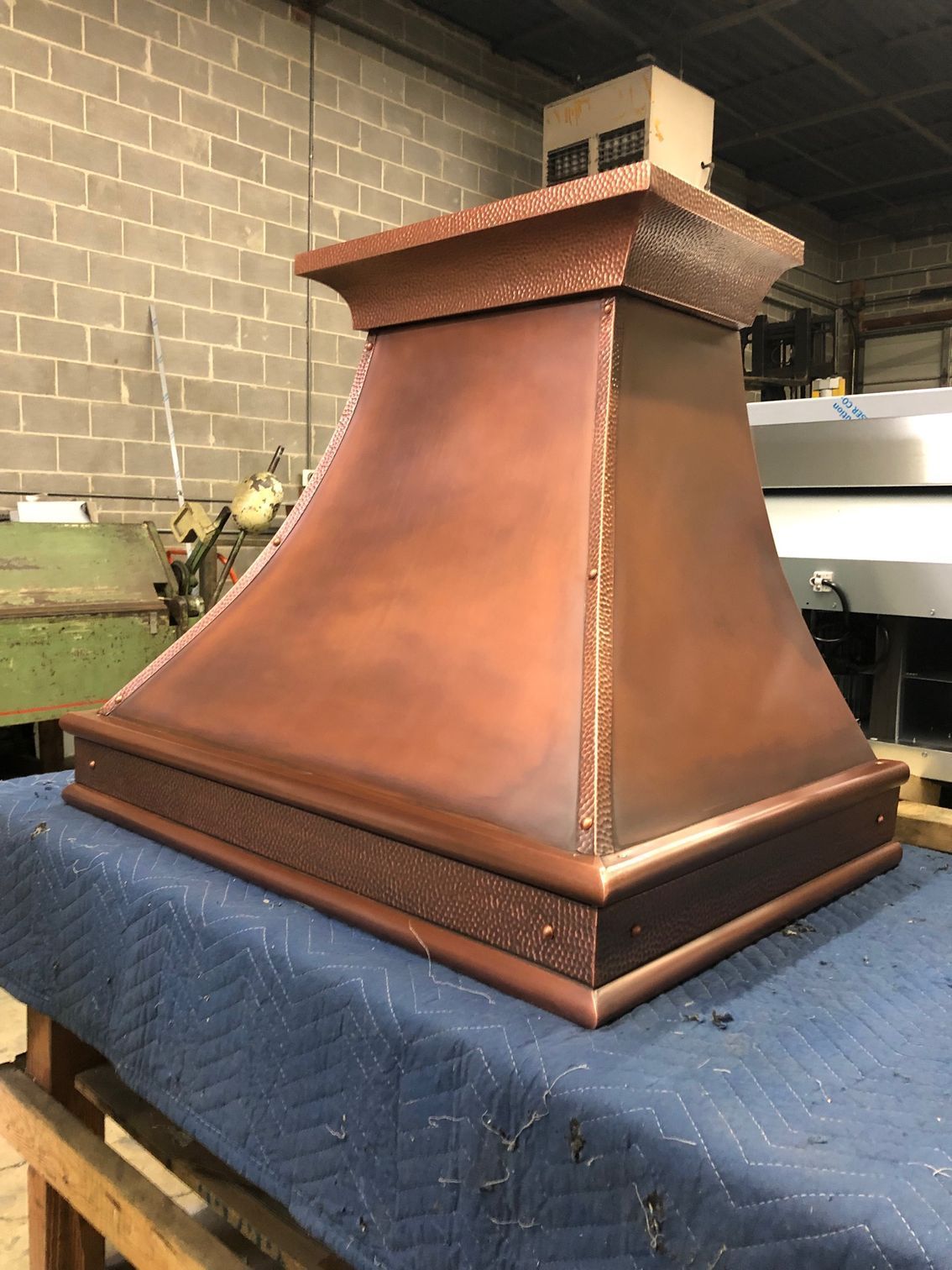 Copper-colored metal range hood with a flared chimney, sitting on a workbench in a workshop.