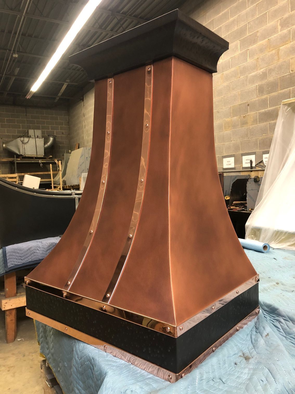 Copper hooded chimney or vent in a workshop, with black trim on a blue-covered table.