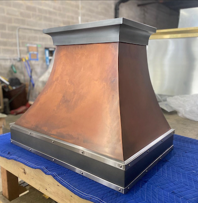 Copper-colored metal chimney hood with a black base, sitting on a blue quilted blanket in a workshop.