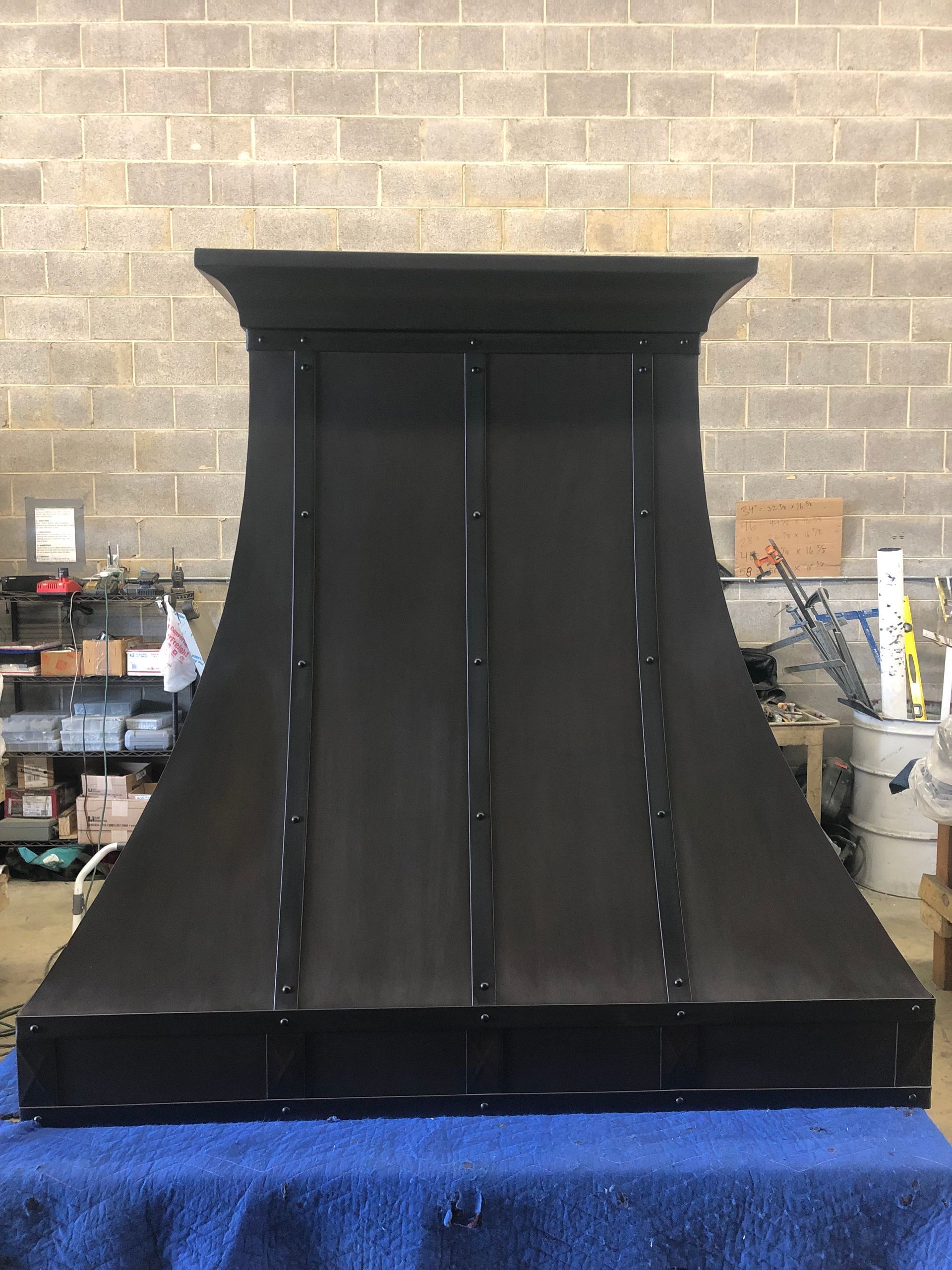 Black metal hood or enclosure standing in a workshop against a brick wall.