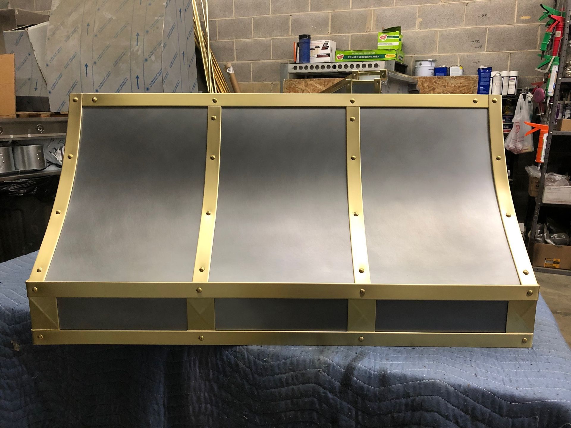 Stainless steel three-bay skate bowl ramp in a workshop, with black riding surfaces and yellow supports.