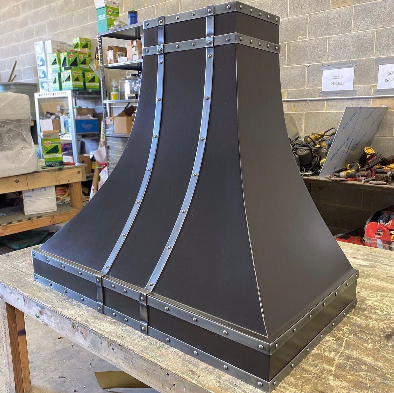 Black metal industrial vent hood in a workshop, with riveted seams and a flared base.