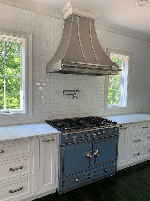 White kitchen with a blue range, stainless hood, white cabinets, and two windows above a marble countertop