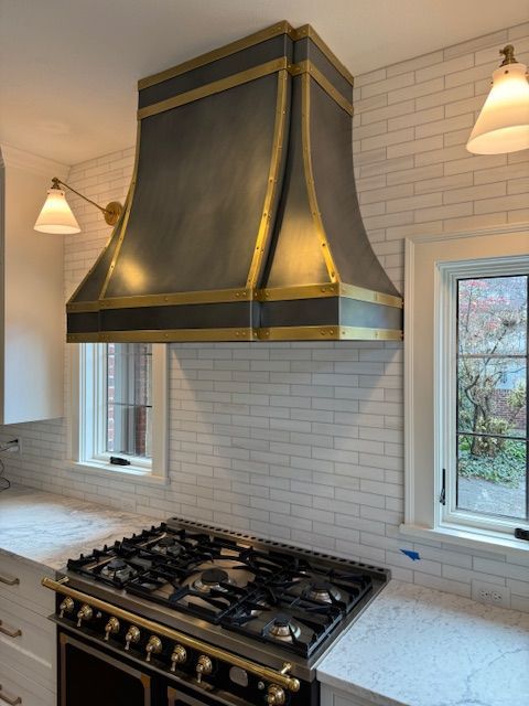 Kitchen stove with brass-accented hood, white tile backsplash, and two pendant lights
