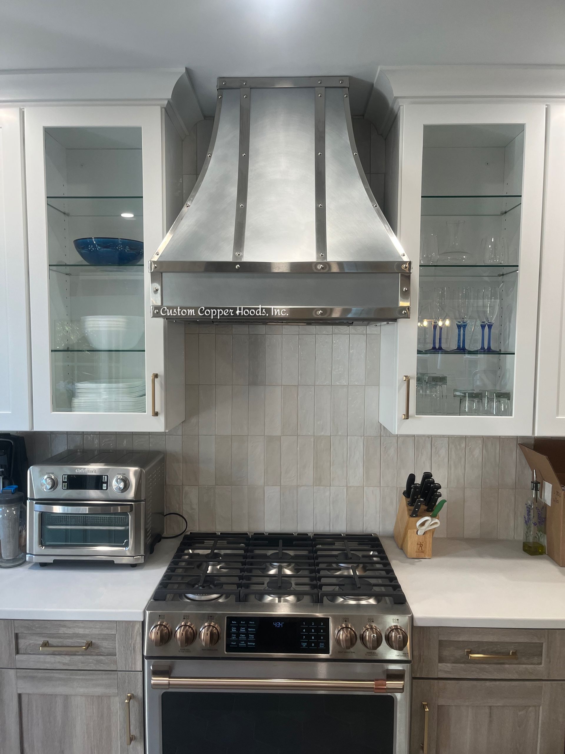 Modern kitchen with stainless steel range hood, gas stove, white cabinets, and glass-front display cabinets.