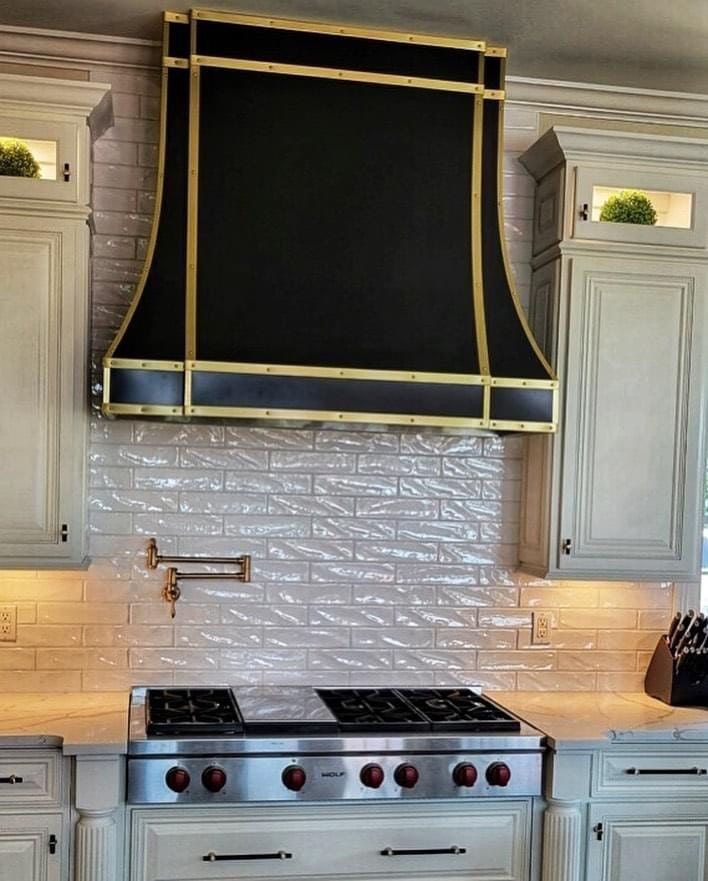 Kitchen stove with a large black-and-gold range hood, white cabinets, and subway tile backsplash.