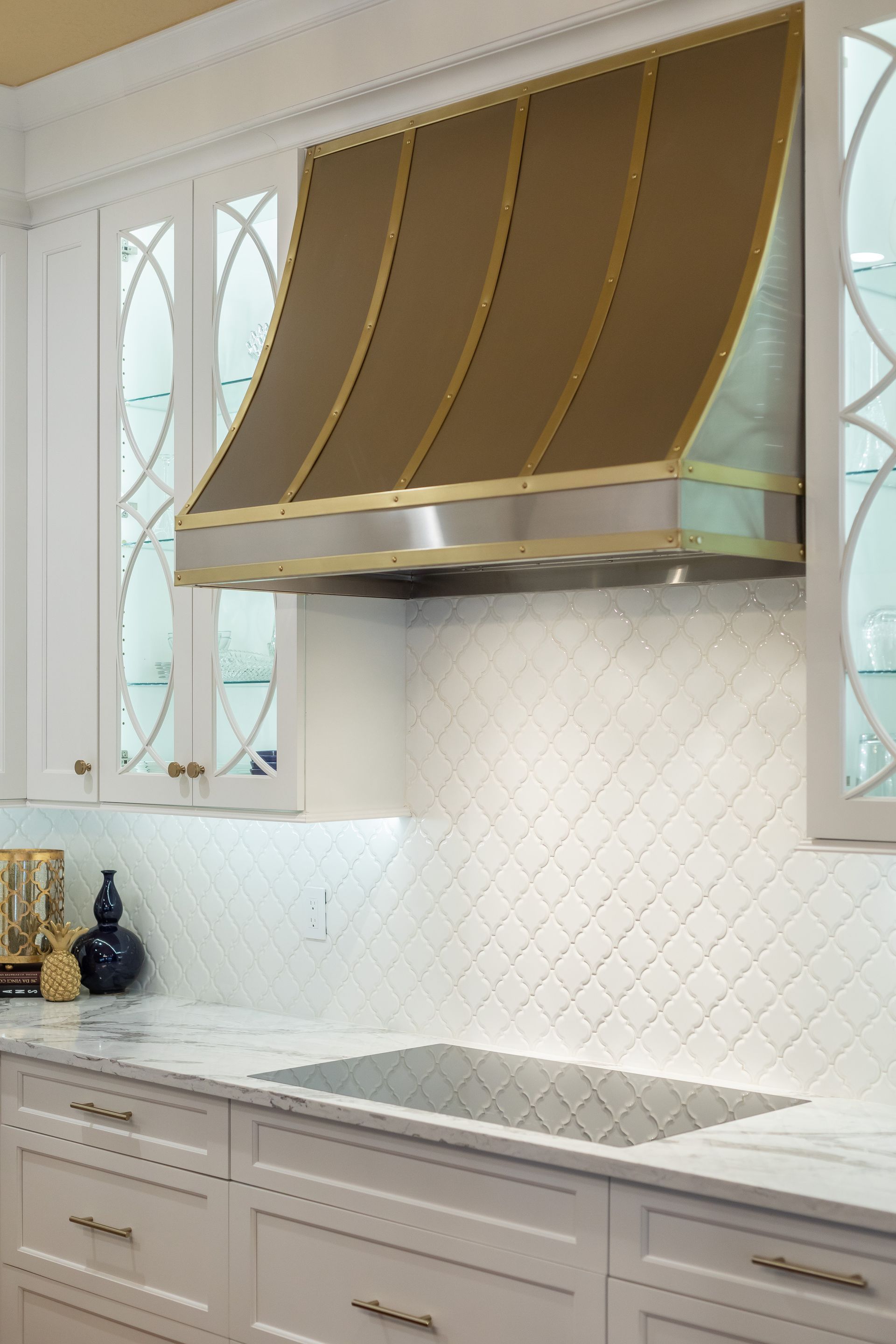 Elegant white kitchen with a gold range hood, marble backsplash, and built-in cooktop