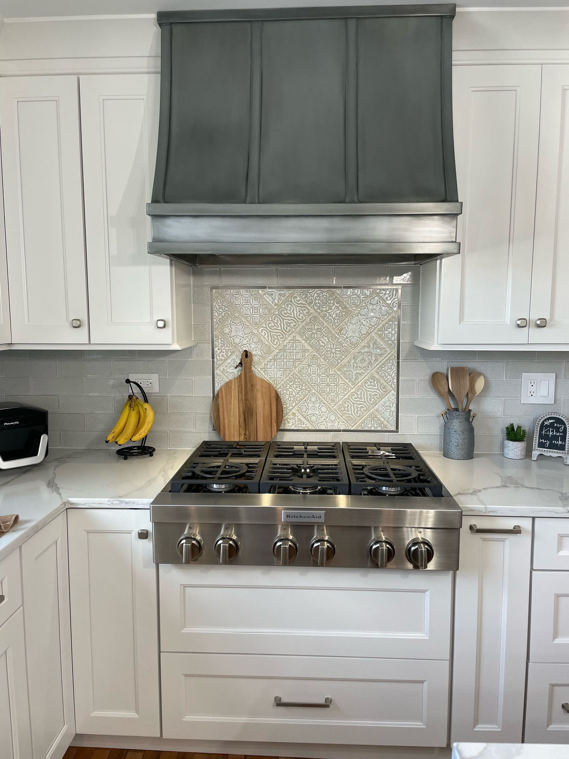 Kitchen with white cabinets, gray range hood, stainless gas stove, and bananas on the counter.