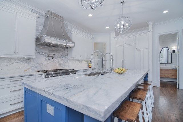 Bright modern kitchen with white marble island, blue cabinets, stainless hood, and bar stools